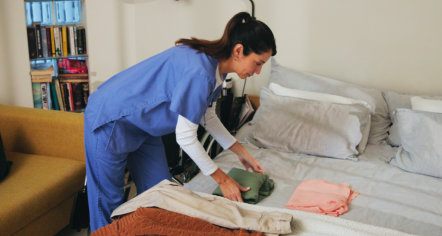 A caregiver in blue scrubs leans over a patient’s bed, adjusting the bedding and a green item; the tidy room feels calm and attentive