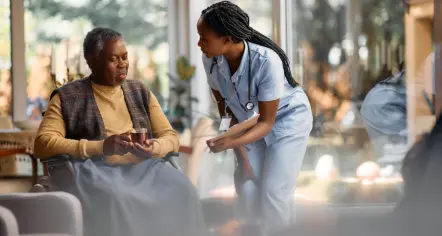 A nurse in blue scrubs leans in to speak with an elderly man seated in a wheelchair holding a cup
