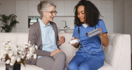 Nurse showing a blood pressure monitor to a smiling senior woman on a couch