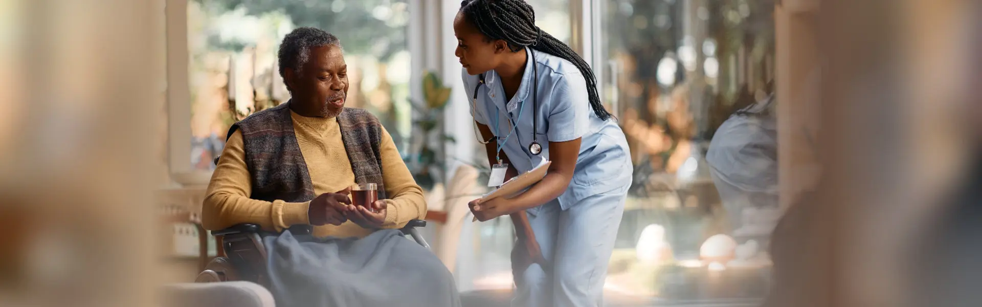 A nurse in blue scrubs leans in to speak with an elderly man seated in a wheelchair holding a cup